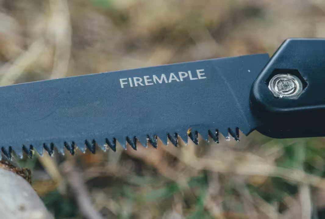 Close-up of a black Firemaple folding knife blade with serrated teeth, showing the brand name 'FIREMAPLE' in white text, set against a blurred outdoor background of dry grass and foliage.