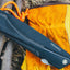 A black Fire Maple folding knife with an orange lanyard, resting on a yellow leather boot against a rustic background of wood and dry grass.