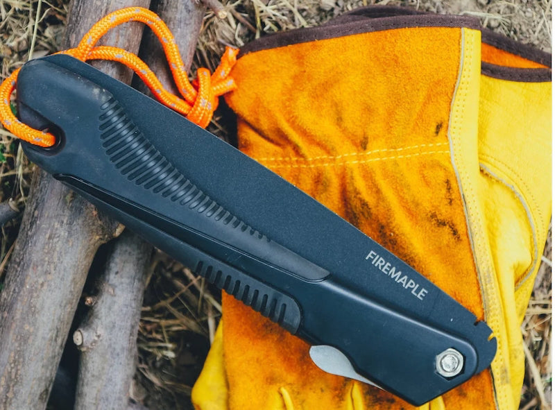 A black Fire Maple folding knife with an orange lanyard, resting on a yellow leather boot against a rustic background of wood and dry grass.