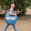 A man in a gray t-shirt, blue jeans, and tan boots walks outdoors carrying a large blue circular camping tent bag with printed white labels.