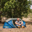 Man sitting next to a blue tent in a natural setting with trees and open space.
