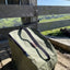 An olive green insulated cooler bag with black zipper and strap details, resting on a weathered wooden fence outdoors with a scenic green hillside in the background.