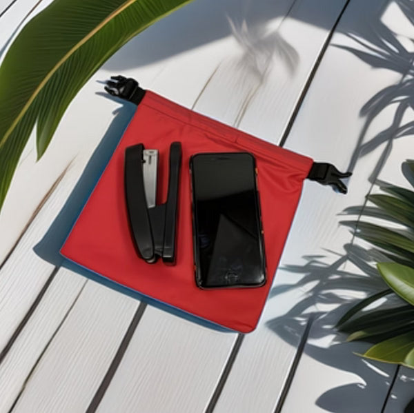 Red pouch with black stapler and phone on a light wooden surface with green leaves.